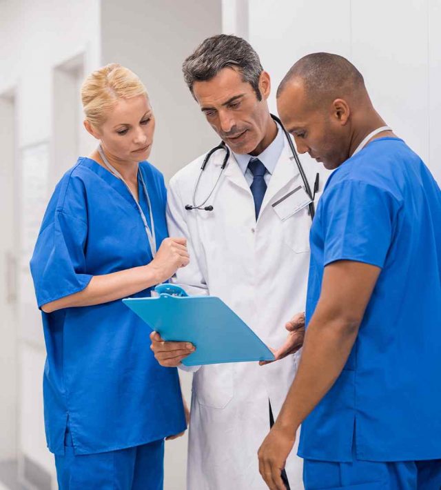 Three health care professionals standing all reviewing a medical record together, two with blue scrubs on and one with a white lab jacket on
