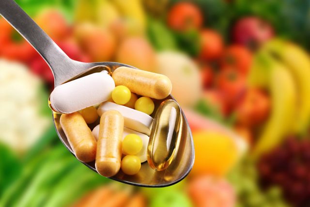 spoon filled with various supplements, capsules, tables, in front of a back drop of various fruits and vegetables